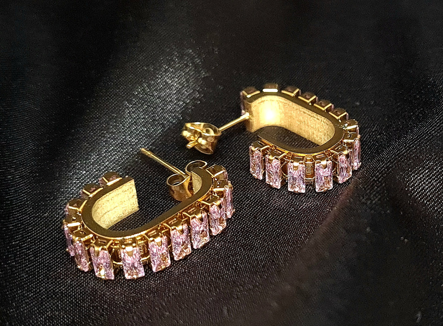 A pair of gold geometric design hoop earrings with pink cubic zirconia stones on a black cloth.