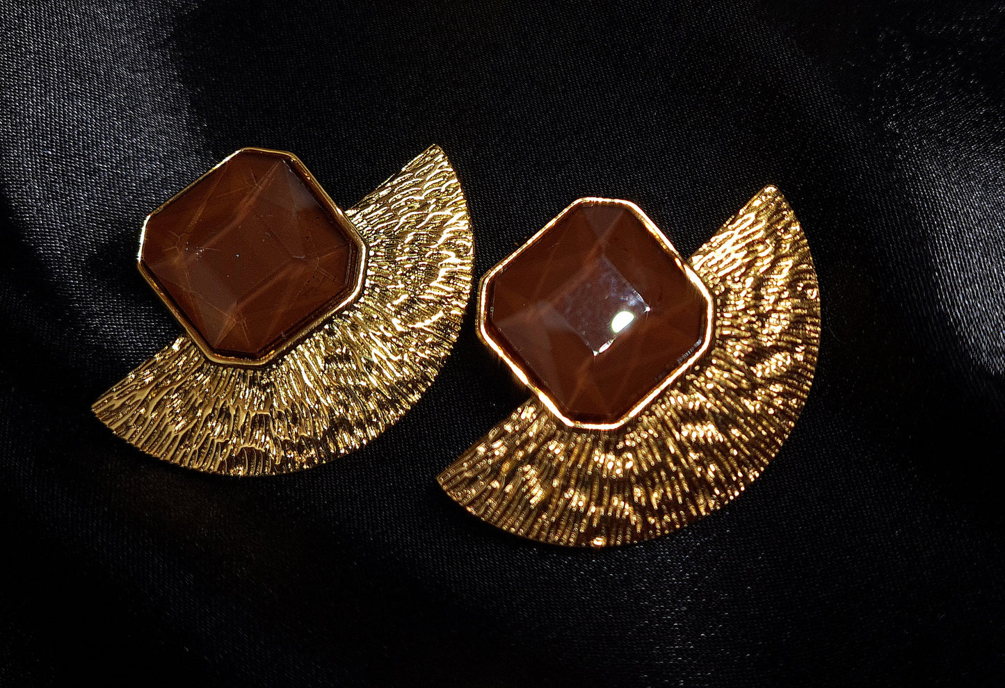 A pair of gold earrings with brown stones on a black surface. The earrings are fan-shaped with a delicate design. They are made of gold with brown stones in the center. The earrings are displayed on a black cloth.