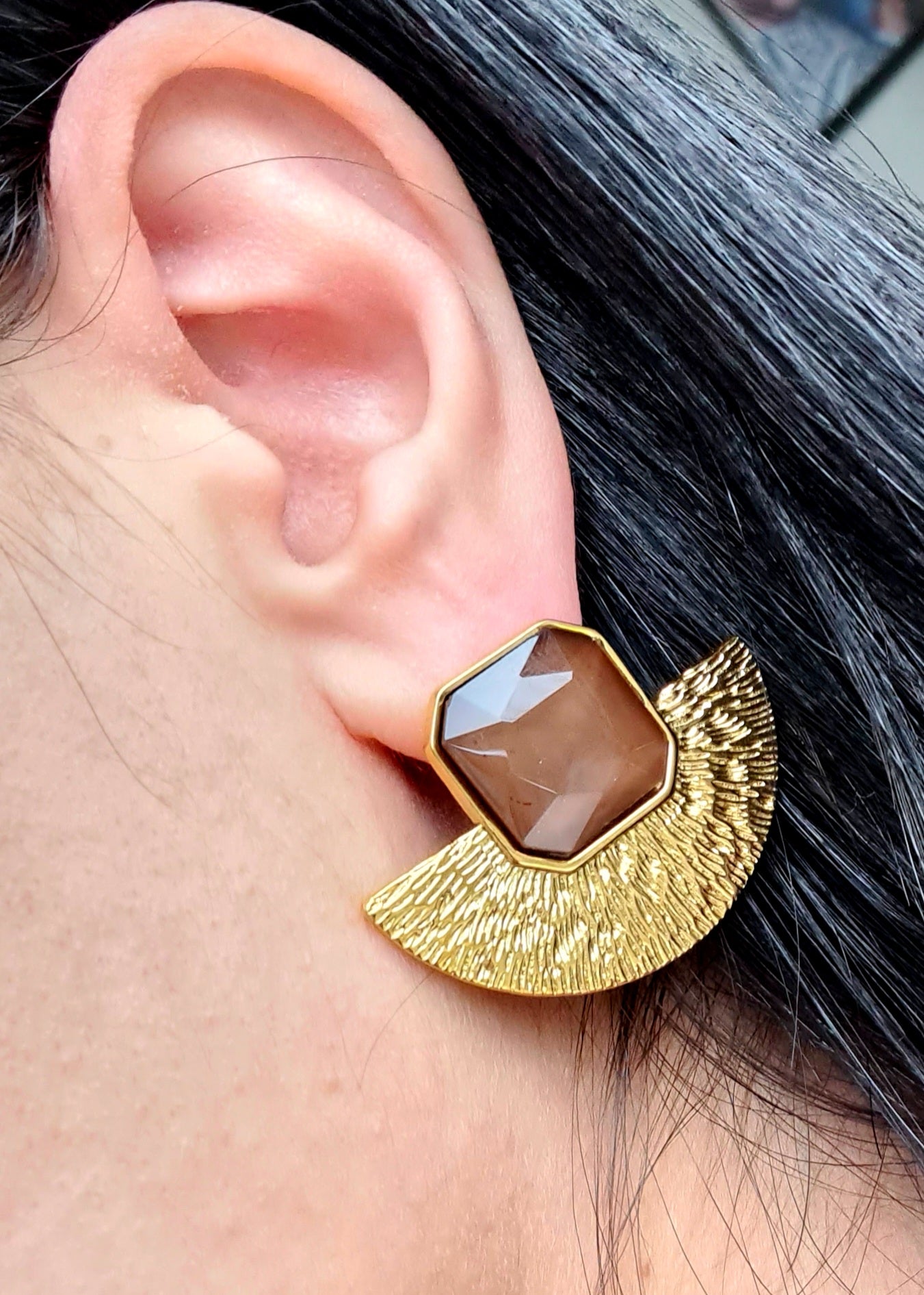 a model wearing A pair of gold earrings with brown stones on a black surface. The earrings are fan-shaped with a delicate design. They are made of gold with brown stones in the center.