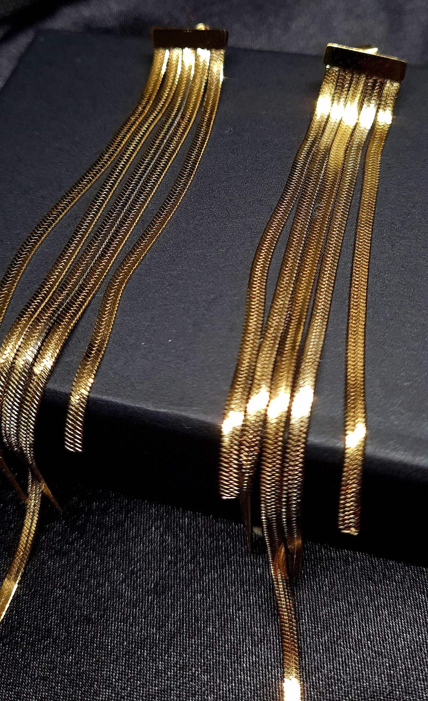 Gold chain earrings sitting on a black box. The earrings are long and have a fringe design. They are made of gold and have a shiny finish. The earrings are sitting on top of a black box.