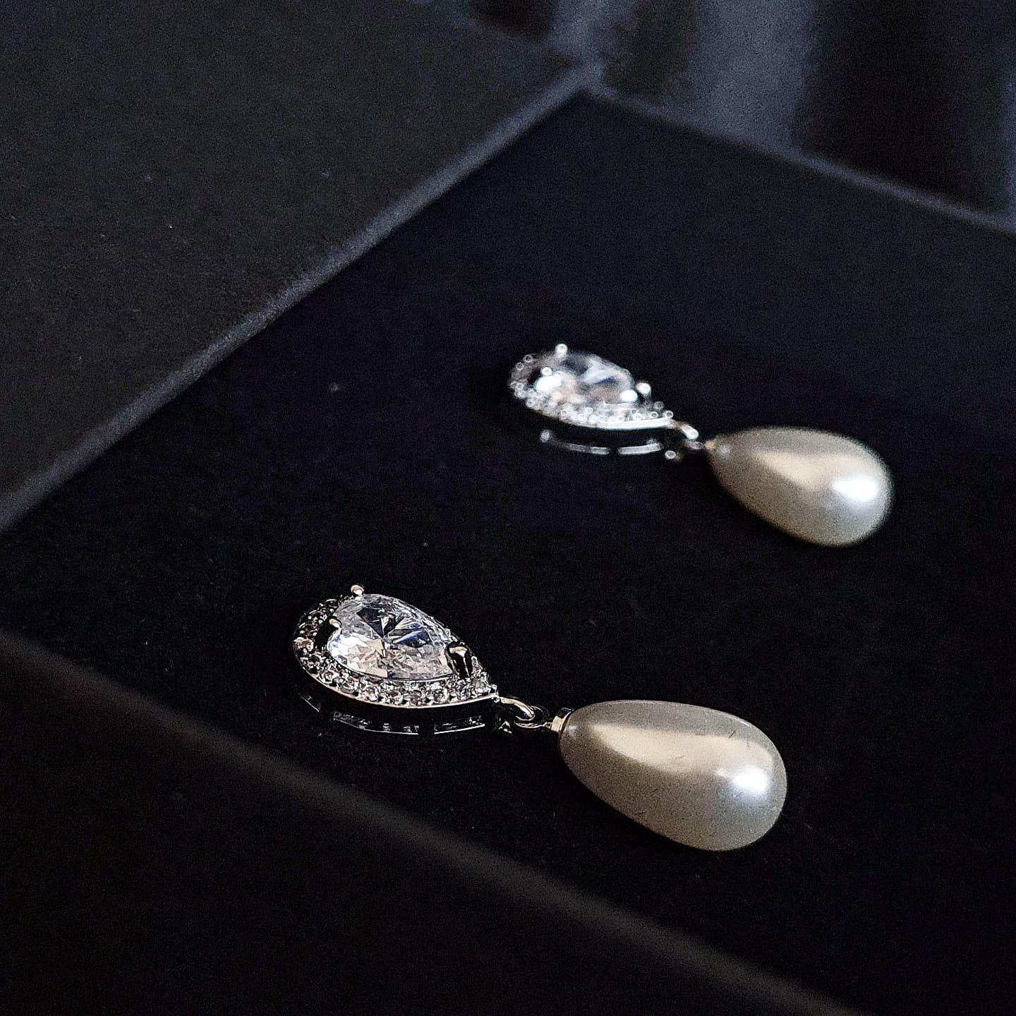 A pair of silver earrings with pearls and diamonds on a black box. The earrings are teardrop-shaped and have a simple, elegant design. The pearls are white and the diamonds are clear.