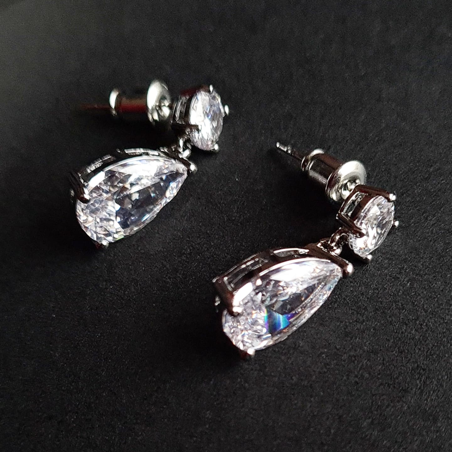 A pair of silver earrings with diamond studs on a black surface. The earrings are teardrop-shaped and have a simple, elegant design. The diamonds are clear and sparkling.