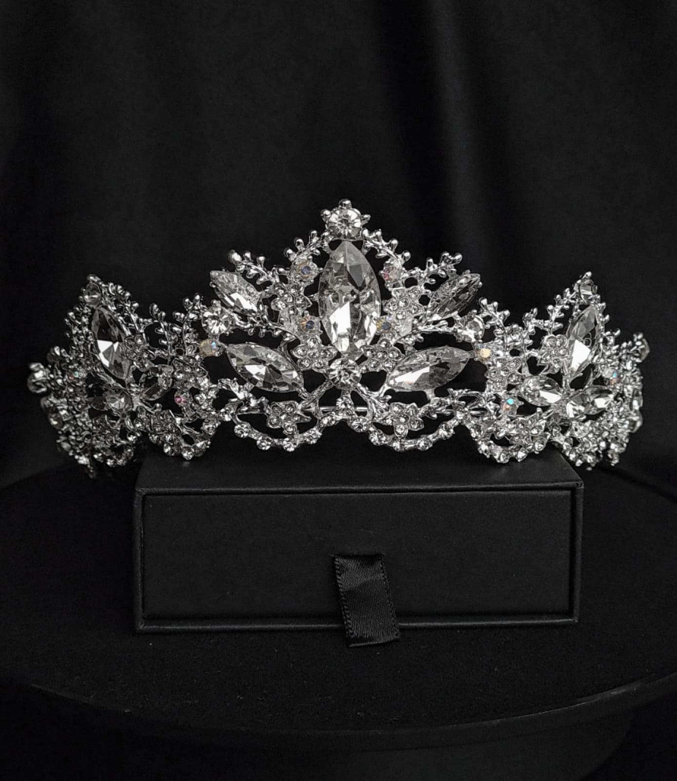 A tiara with rhinestones on a black box. The tiara is made of silver and features a delicate design with cascading rhinestones. The rhinestones are clear and sparkling. The tiara is sitting on top of a black box.