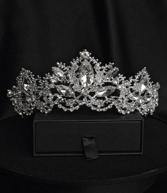 A tiara with rhinestones on a black box. The tiara is made of silver and features a delicate design with cascading rhinestones. The rhinestones are clear and sparkling. The tiara is sitting on top of a black box.