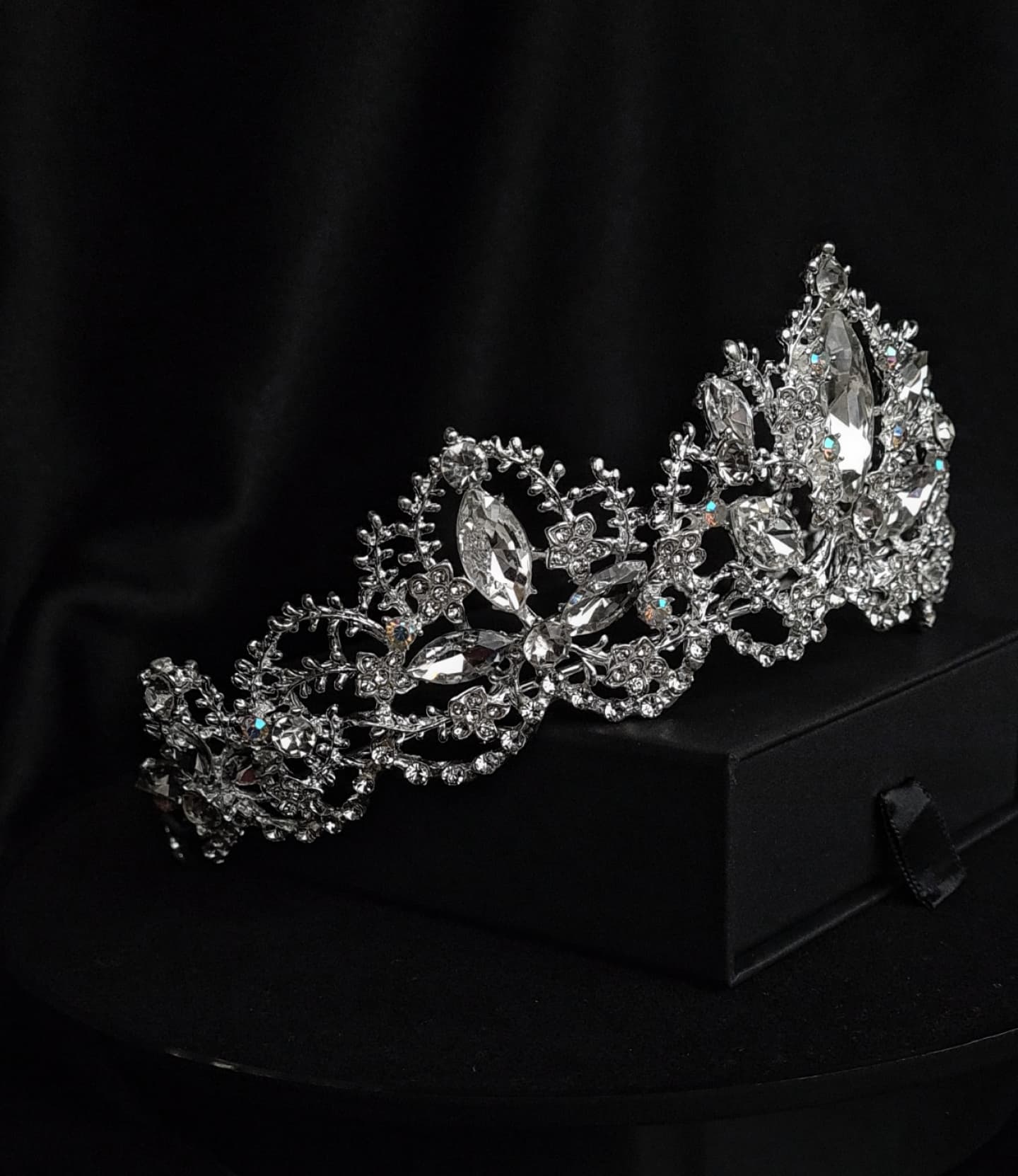 A tiara with rhinestones on a black box. The tiara is made of silver and features a delicate design with cascading rhinestones. The rhinestones are clear and sparkling. The tiara is sitting on top of a black box.