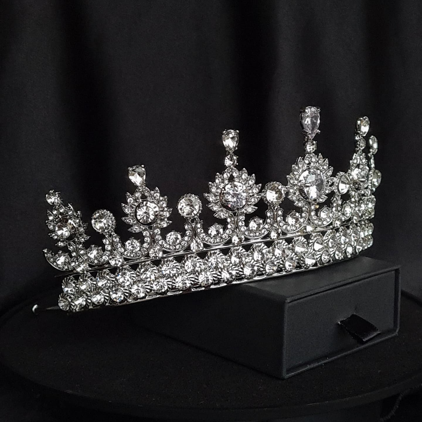 A close-up of a tiara with rhinestones on a black background. The tiara is made of silver and features a delicate design with cascading rhinestones. The rhinestones are clear and sparkling.
