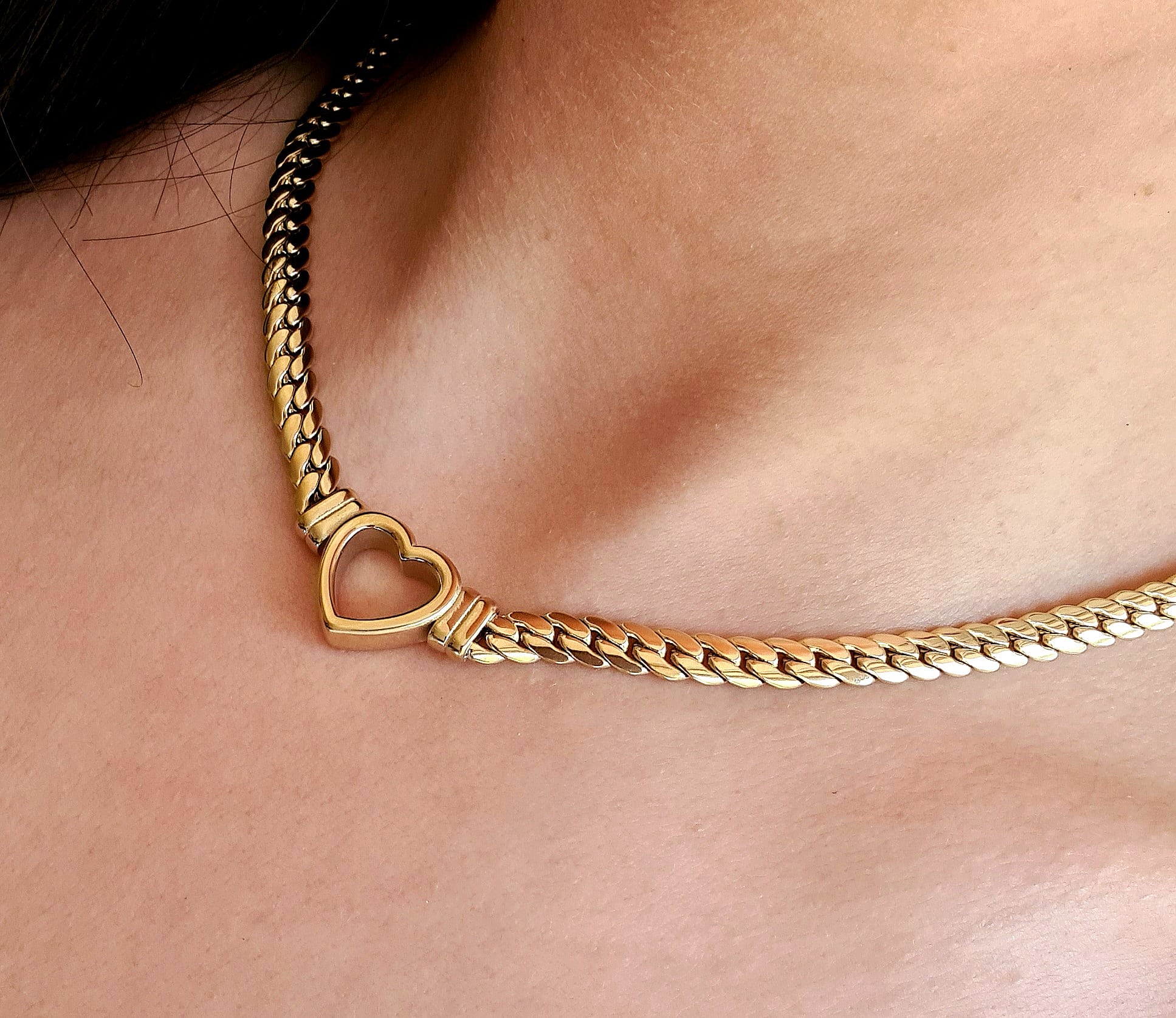 Woman wearing a Gold necklace with heart pendant, the necklace is made of gold and has a heart pendant in the center. The heart is gold and has a smooth surface.