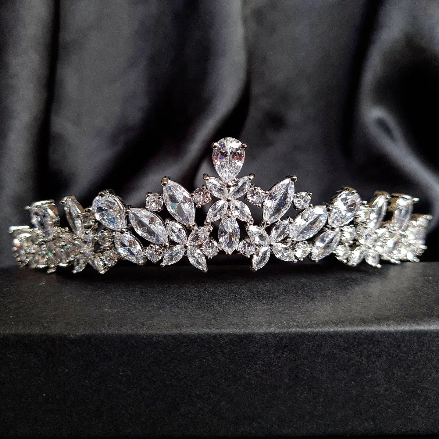 A tiara with diamonds on a black background. The tiara is made of silver and features a delicate design with cascading diamonds. The diamonds are clear and sparkling. The tiara is sitting on a table.
