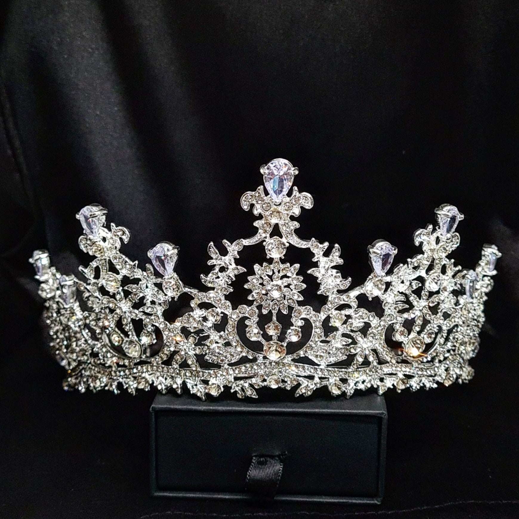 A silver tiara with rhinestones and flowers sitting on a black box. The tiara is made up of multiple curved bands, each of which is decorated with rhinestones and flowers. The tiara is sitting on top of a black box, which is partially obscuring the bottom of the tiara.