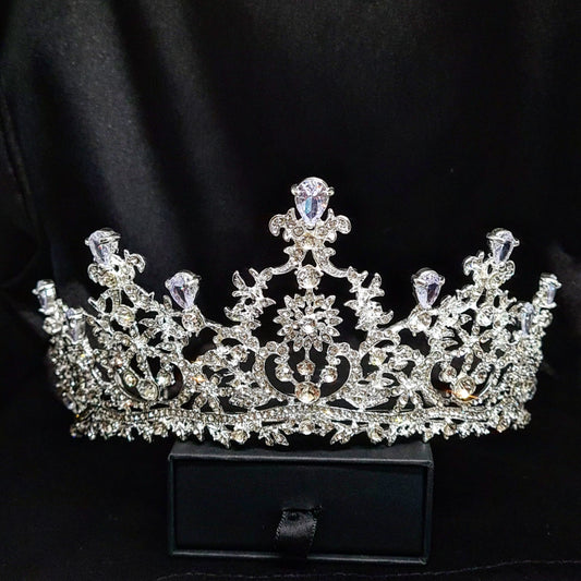 A silver tiara with rhinestones and flowers sitting on a black box. The tiara is made up of multiple curved bands, each of which is decorated with rhinestones and flowers. The tiara is sitting on top of a black box, which is partially obscuring the bottom of the tiara.