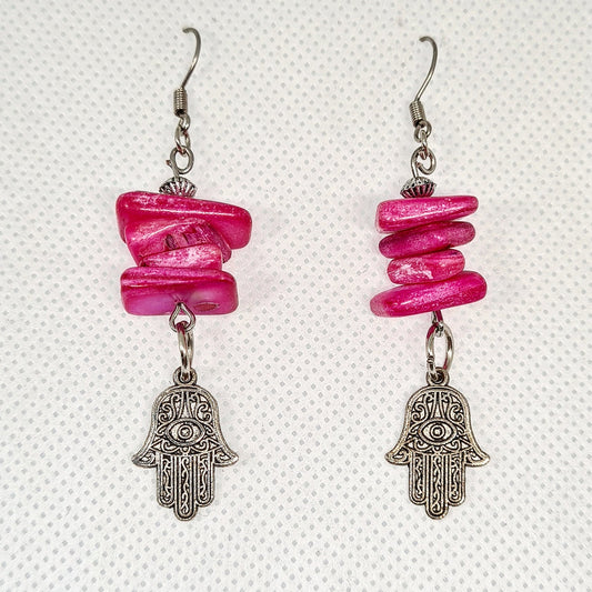 Pair of pink earrings with hamsa pendant. The earrings are made of pink metal and have a hamsa pendant in the center. The hamsa is a hand-shaped amulet that is believed to bring good luck and protection. The earrings are hanging from gold hooks.