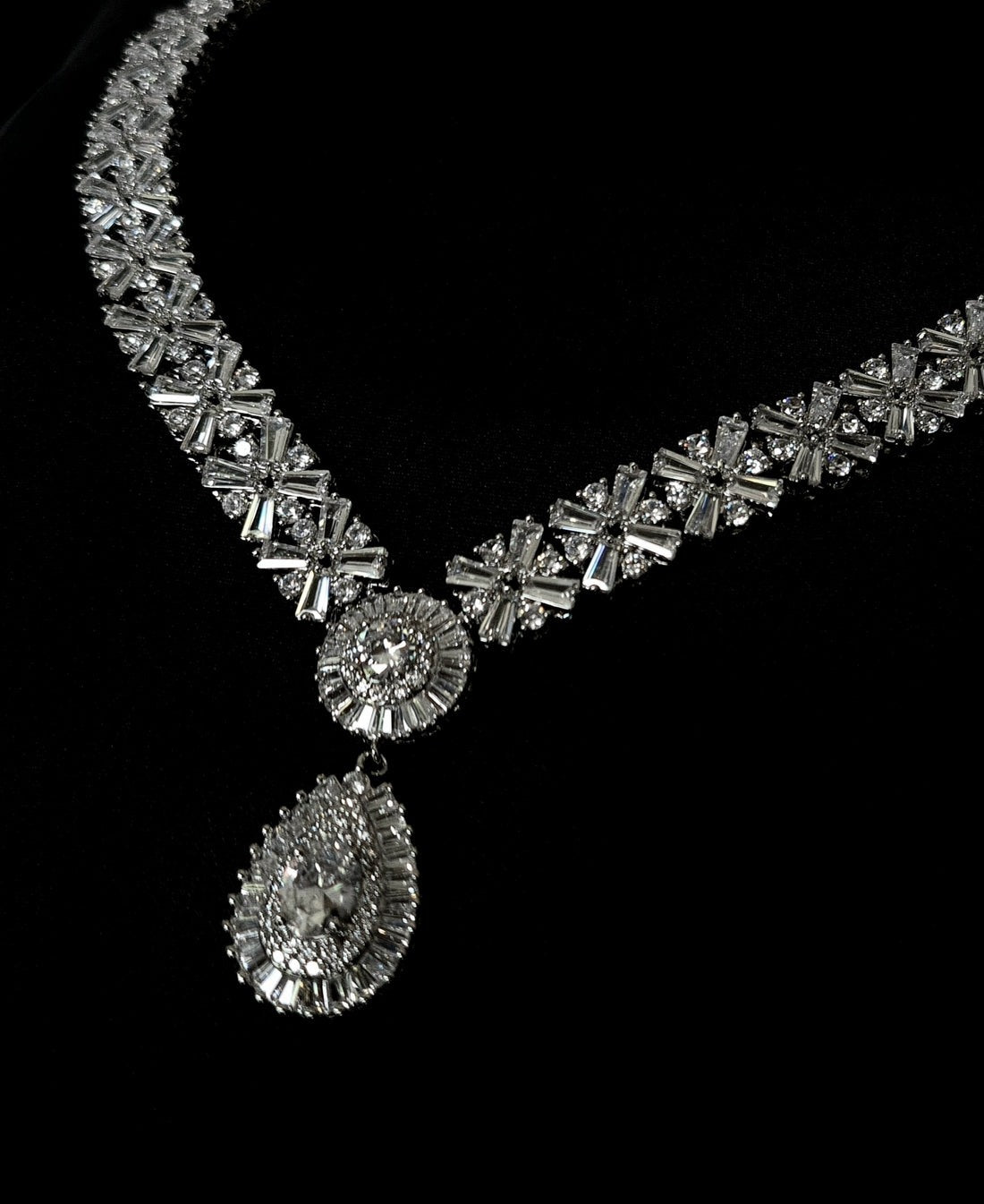a necklace with a large diamond pendant on a black background. The pendant is round and has a white diamond in the center. The necklace is made of silver and has a big chain. The background is black.