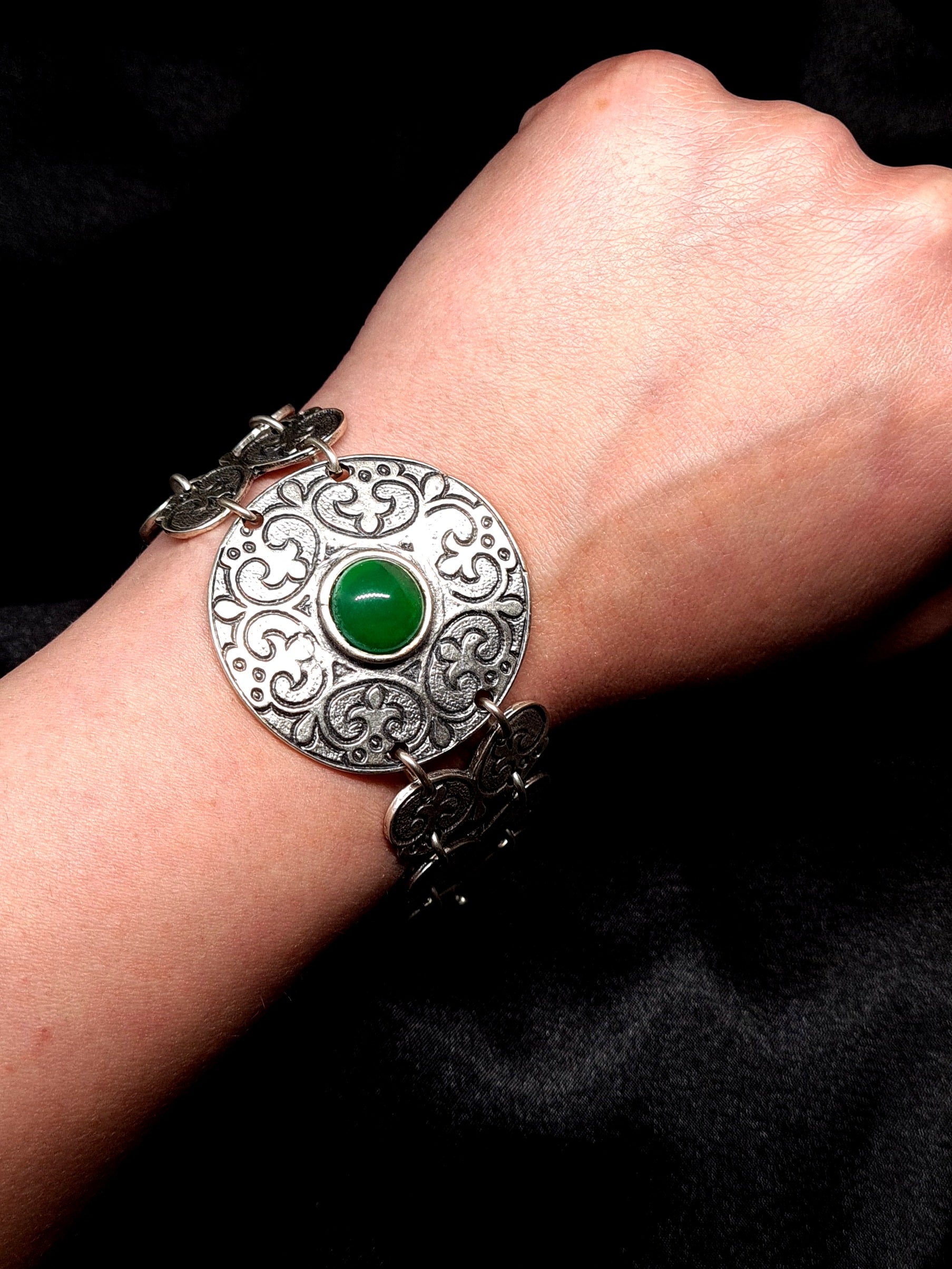 A silver bracelet with a green stone in the center. The bracelet is made of silver and has a delicate design. The green stone is oval-shaped and has a smooth finish. The bracelet is on a woman's wrist.