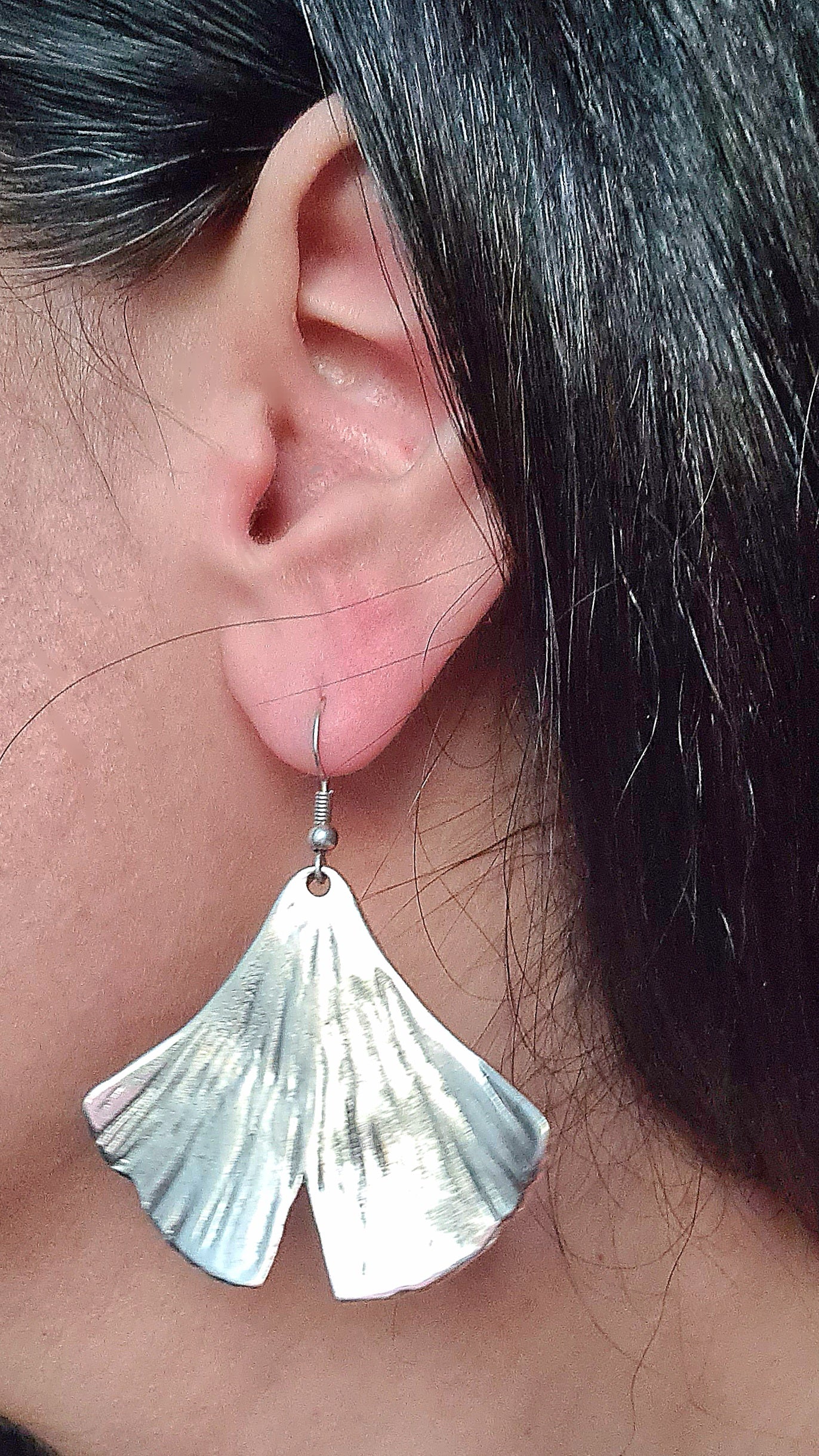 a woman wearing a pair of silver earrings in the shape of a ginkgo leaf . The earrings are small and delicate, and they have a shiny finish. The ginkgo leaf is fan-shaped and has a textured appearance.