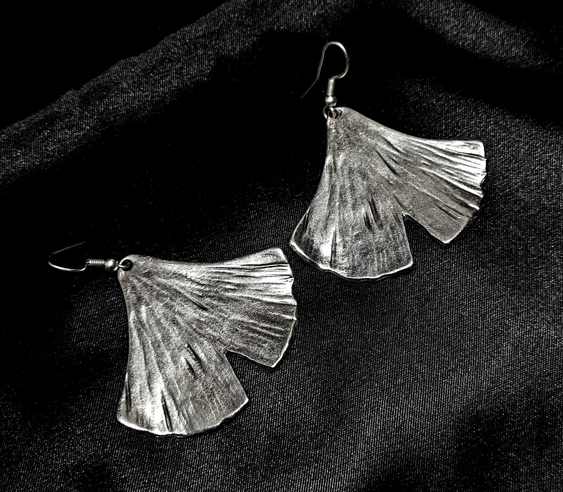 A pair of silver earrings in the shape of a ginkgo leaf sitting on a black surface. The earrings are small and delicate, and they have a shiny finish. The ginkgo leaf is fan-shaped and has a textured appearance.