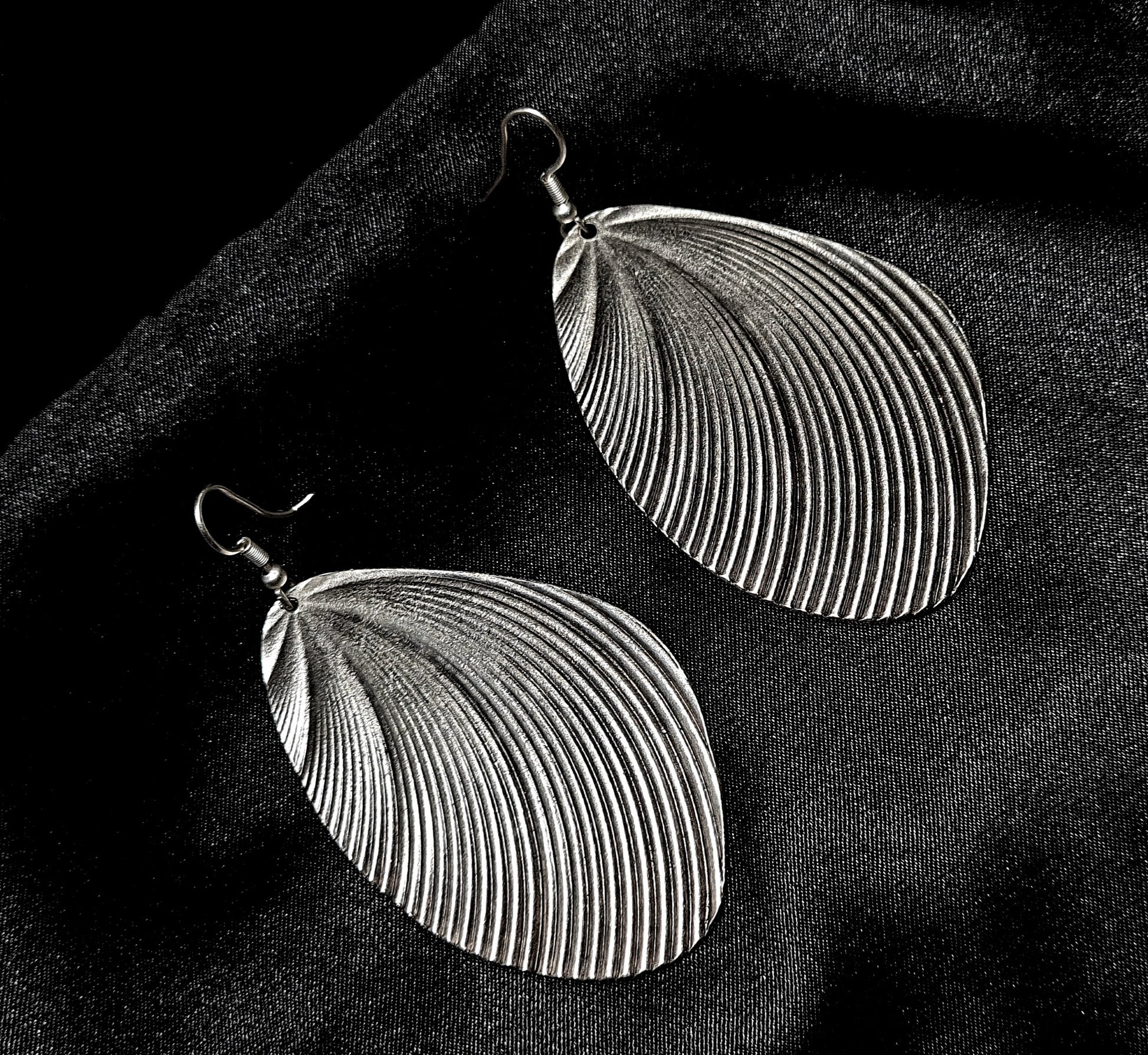 A pair of silver earrings with a shell design sitting on a black cloth. The earrings are small and delicate, and they have a shiny finish. The shell design is in the shape of a seashell, and it has a textured appearance