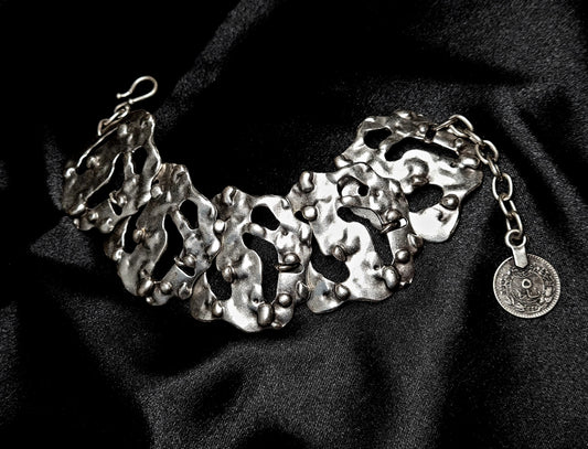 A silver bracelet sitting on a black cloth. The bracelet is made of silver and has a delicate design. The coin is small and round, and it has a silver color. The bracelet is on a black cloth.