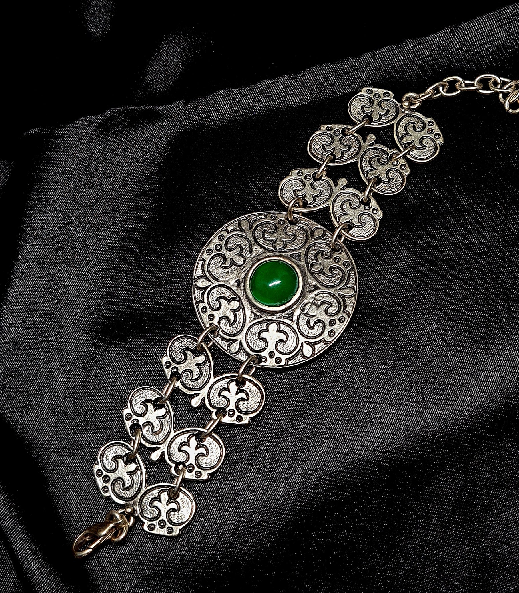 A silver bracelet with a green stone in the center. The bracelet is made of silver and has a delicate design. The green stone is oval-shaped and has a smooth finish. 