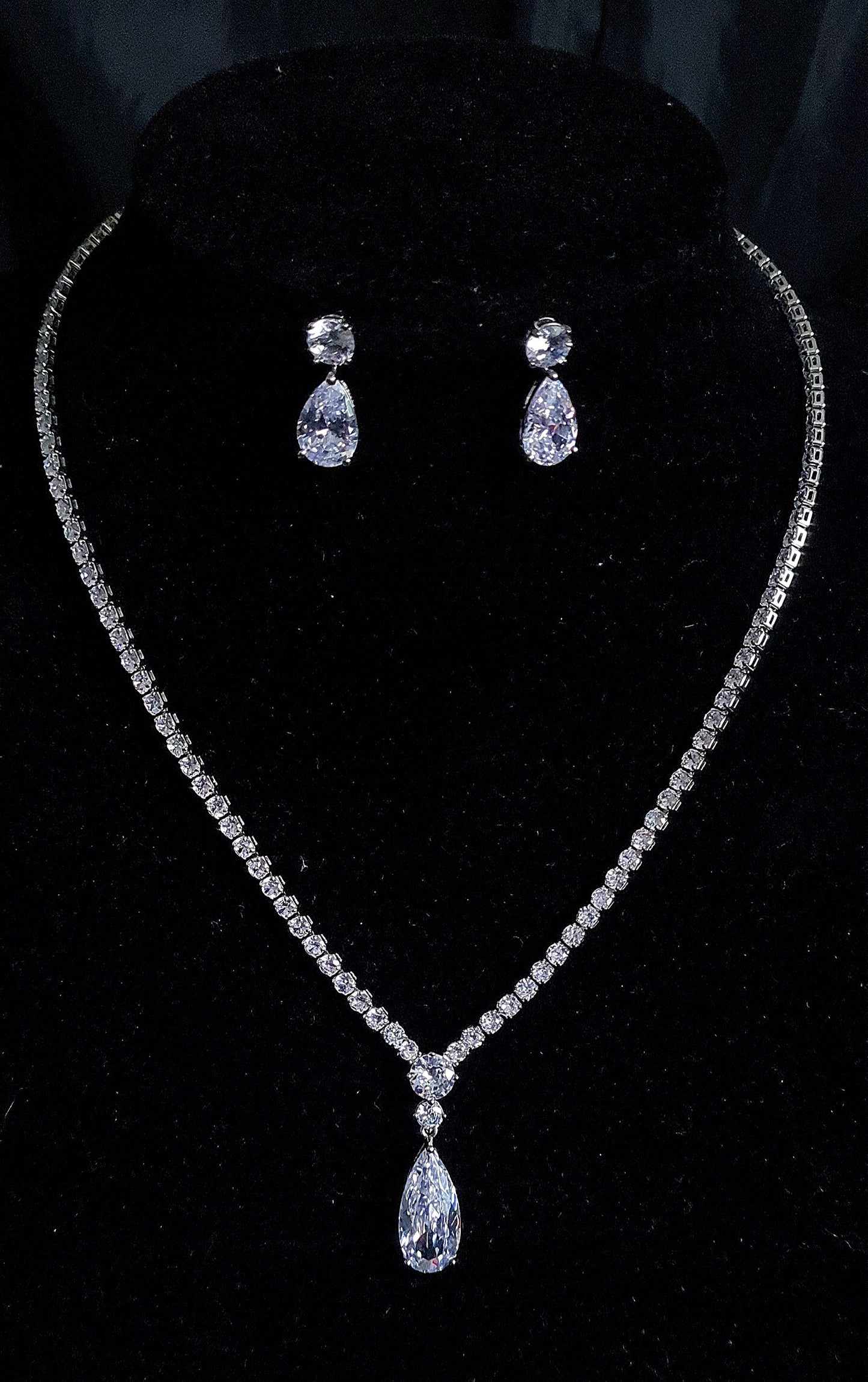 A set of sparkling necklace and earrings on a black surface. The necklace is long and flowing with a delicate design. It is made of silver with sparkling cubic zirconia crystals. The earrings are matching teardrop shapes with a similar design. The necklace and earrings are displayed on a black surface. The background is also black.