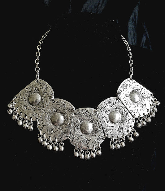 A silver necklace with a large oval-shaped pendant. The pendant is made of silver and has a delicate design. The pendant is decorated with small round stones in a silver color. The necklace is long and hangs down to the chest.