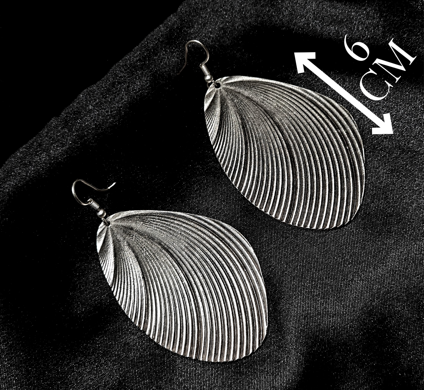 A pair of silver earrings with a shell design sitting on a black cloth. The earrings are small and delicate, and they have a shiny finish. The shell design is in the shape of a seashell, and it has a textured appearance