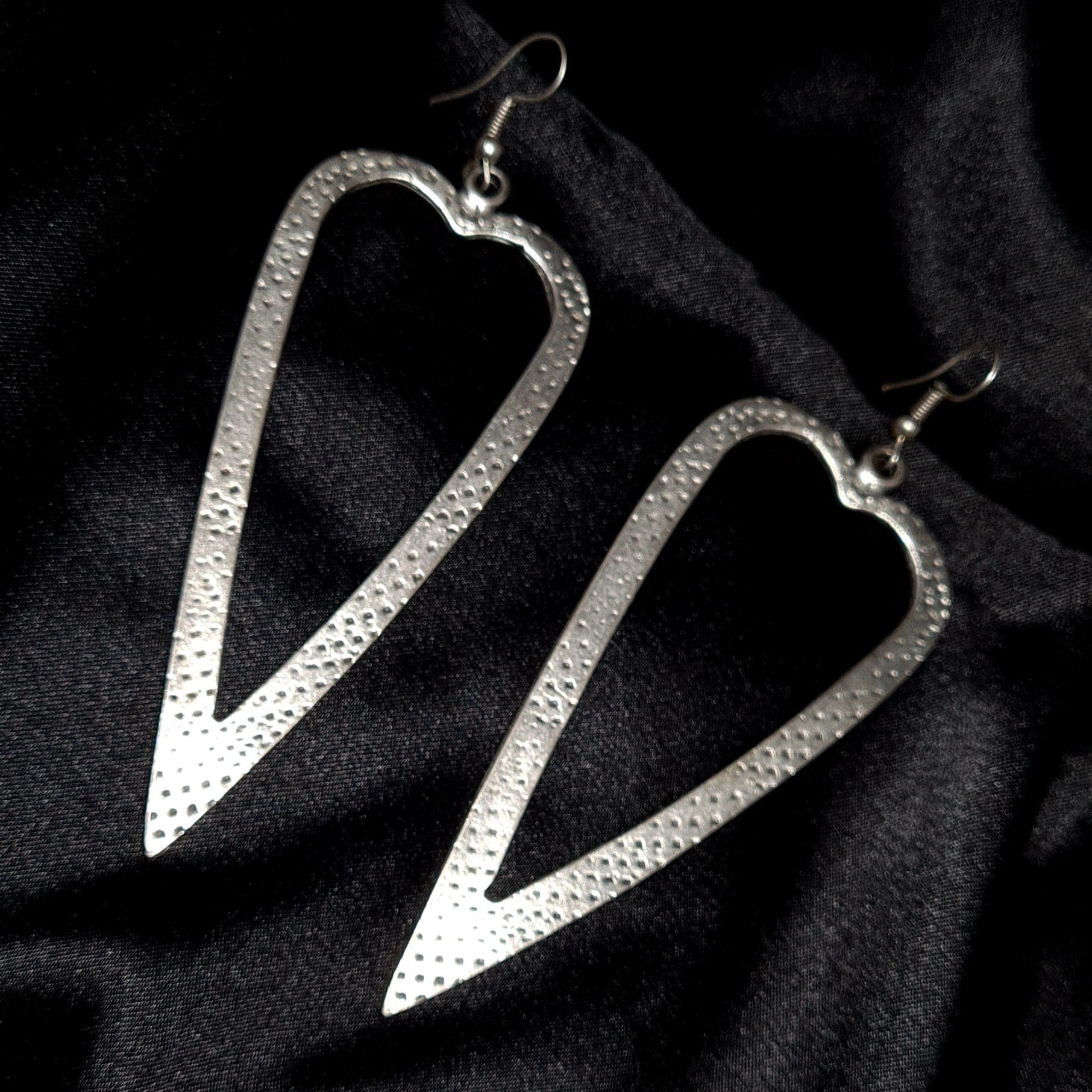 A pair of silver earrings in the shape of a heart on a black background. The earrings are small and delicate, and they have a shiny finish. The heart is smooth and has a rounded shape.