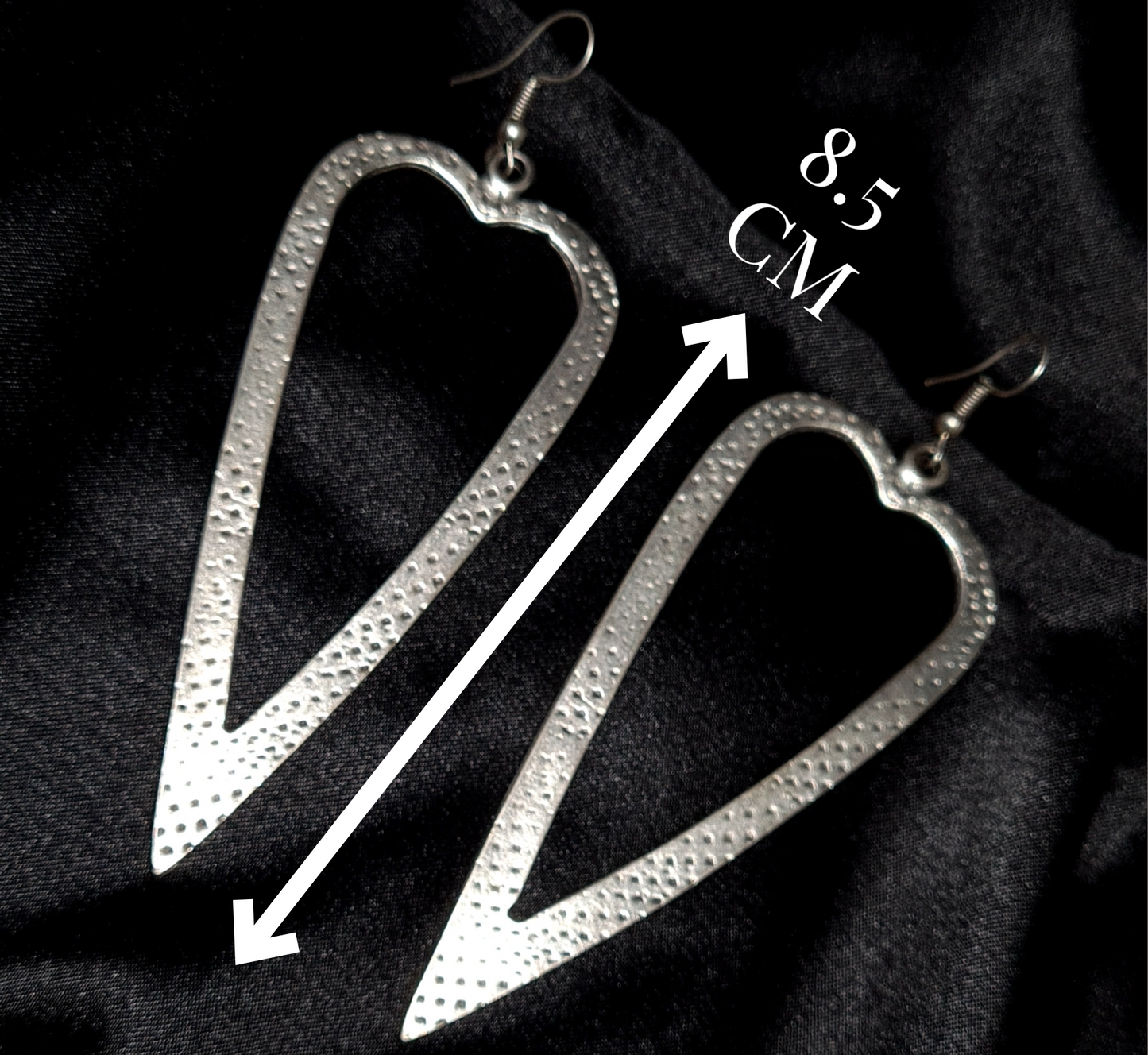 A pair of silver earrings in the shape of a heart on a black background. The earrings are small and delicate, and they have a shiny finish. The heart is smooth and has a rounded shape with measures