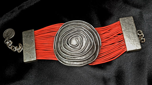 A red bracelet with a silver swirl on it. The bracelet is made of red thread and has a silver swirl design on it. The swirl is big and delicate, and it is in the center of the bracelet. 