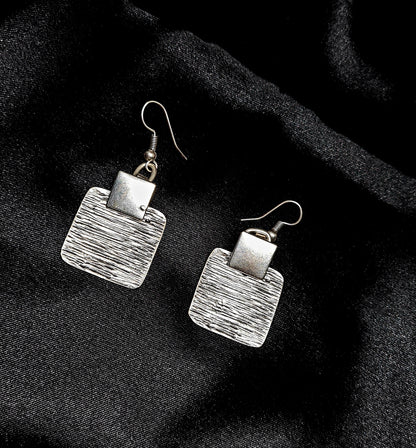 A pair of silver earrings sitting on a black cloth. The earrings are small and delicate, and they have a square shape. The earrings are made of silver and have a shiny finish.