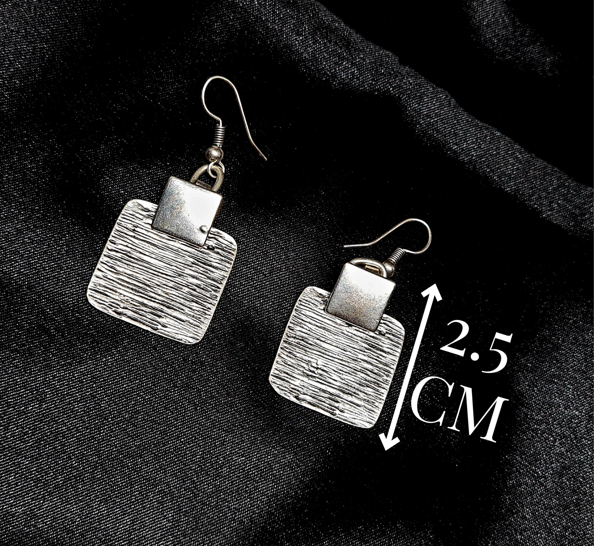 A pair of silver earrings sitting on a black cloth. The earrings are small and delicate, and they have a square shape. The earrings are made of silver and have a shiny finish with measures.