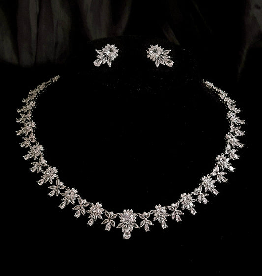 A close-up of a necklace and earrings set. The necklace is a silver chain with a pendant in the shape of a flower. The earrings are princess cut shape. The background is a black velvet cloth.