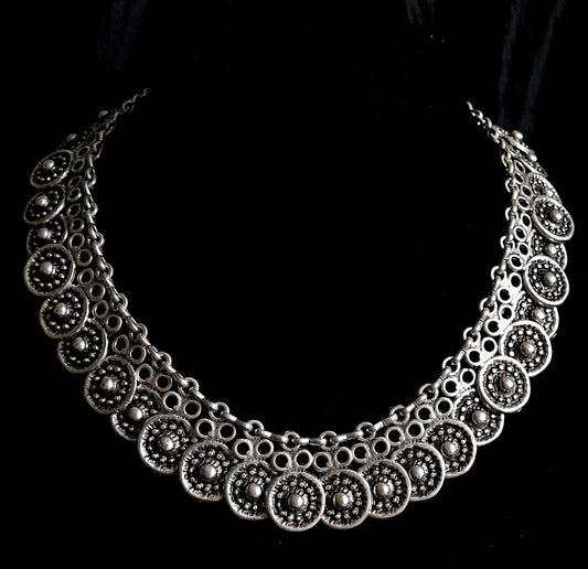 A silver necklace with circles on a black background. The necklace is made of silver and has a delicate design. The circles are small and have a smooth finish. The necklace is long and hangs down to the chest.
