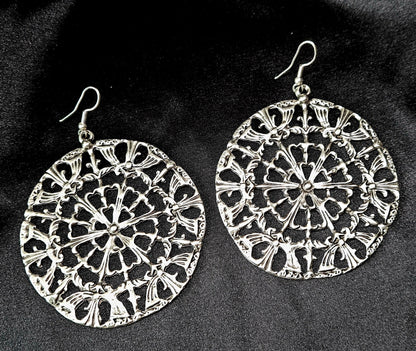 A pair of silver earrings on a black surface. The earrings are made of silver and have a circular design on them. The earrings are small and delicate, and they have a shiny finish.