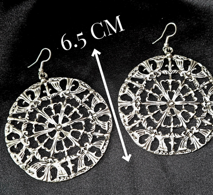 A pair of silver earrings on a black surface. The earrings are made of silver and have a circular design on them. The earrings are small and delicate, and they have a shiny finish with length