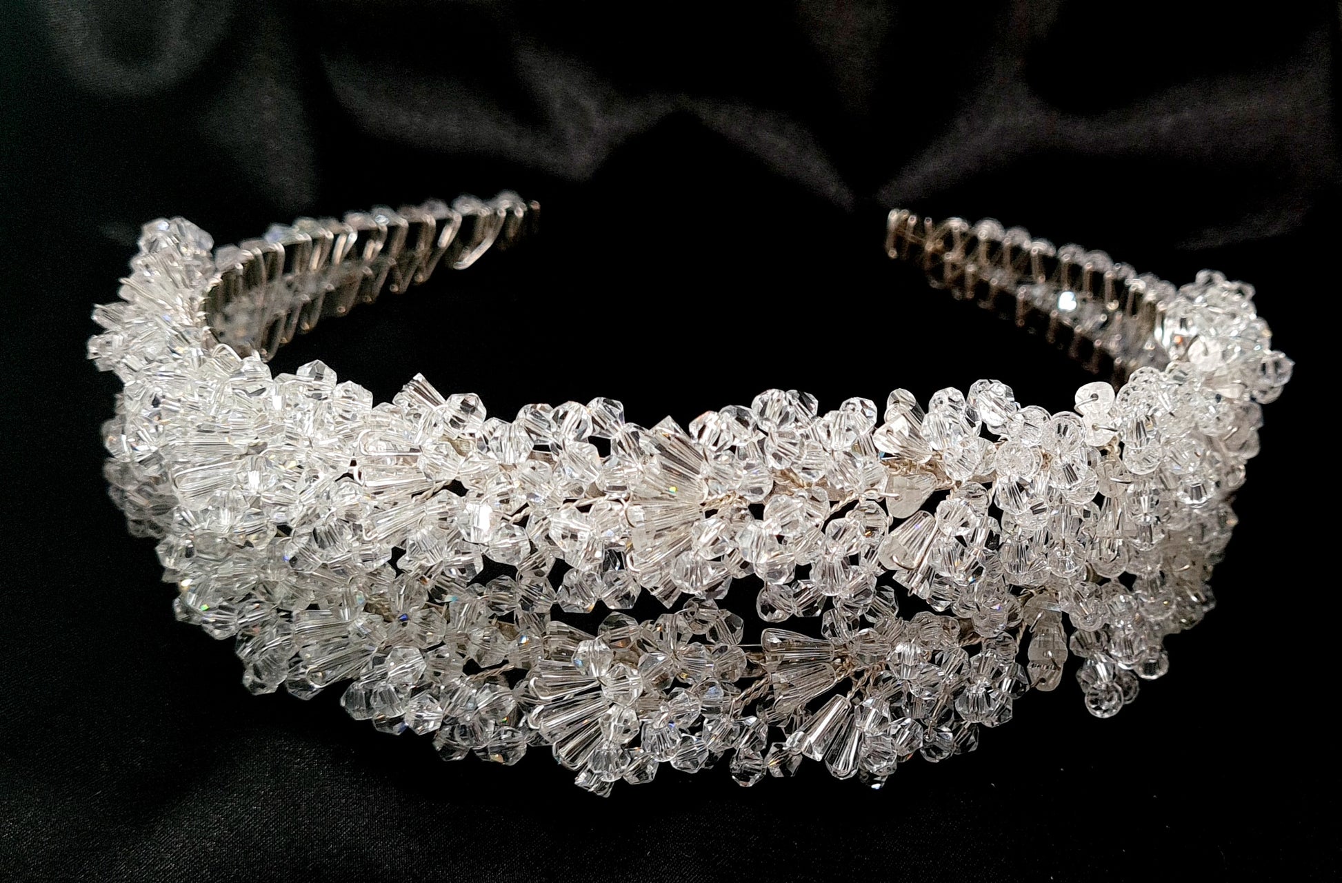 A close-up of a headband with crystals on a black background. The headband is made of a silver-colored metal and has an intricate design with crystals of different shapes and sizes. The crystals are sparkling and reflect the light.