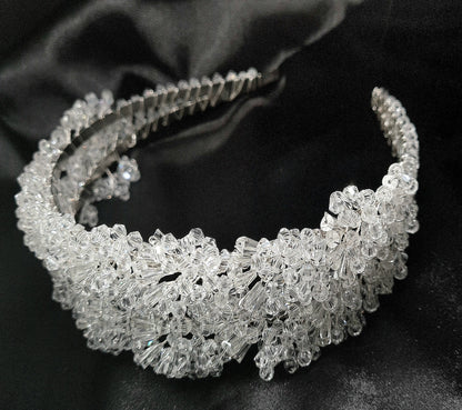 A close-up of a headband with crystals on a black background. The headband is made of a silver-colored metal and has an intricate design with crystals of different shapes and sizes. The crystals are sparkling and reflect the light.