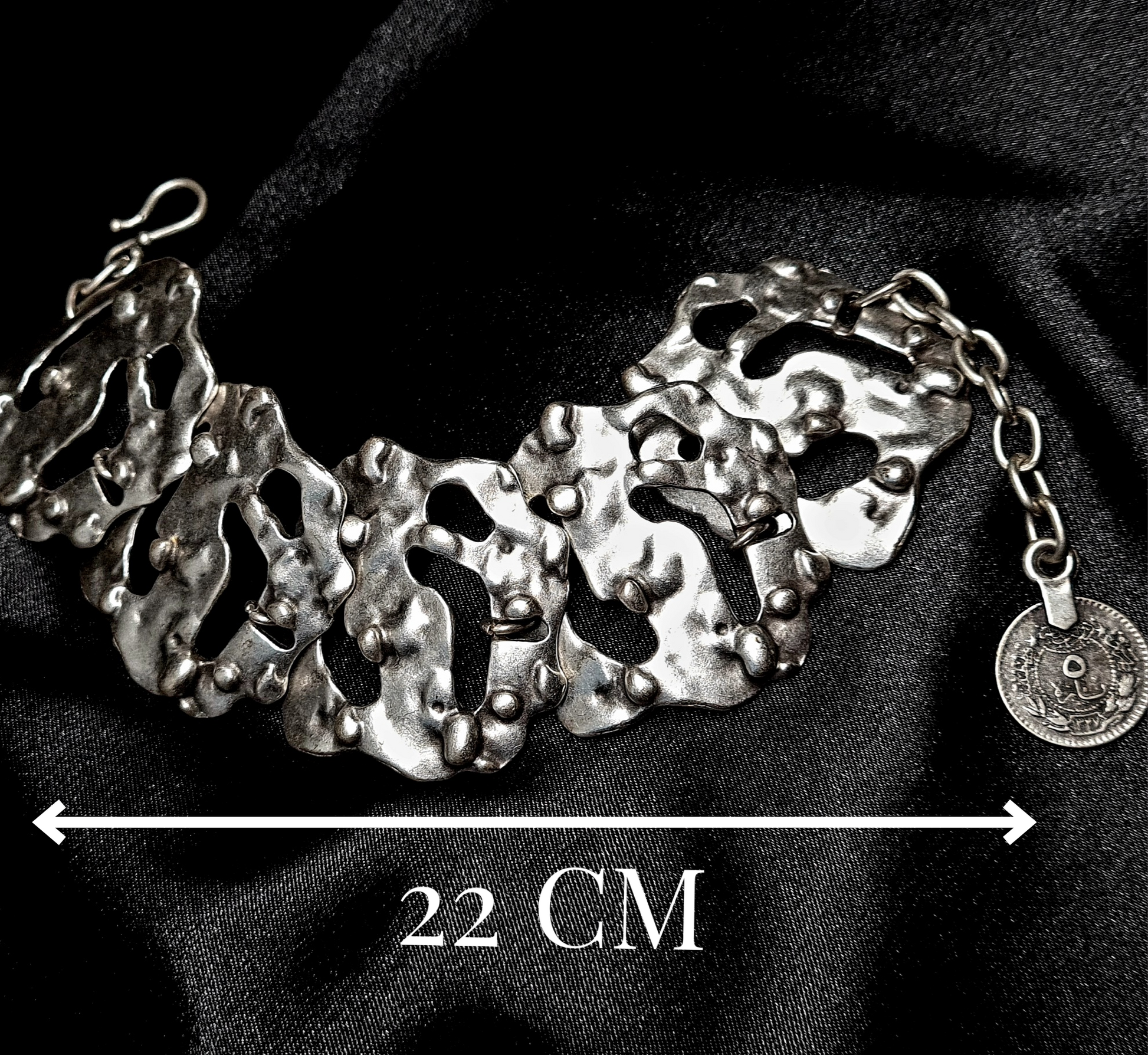 A silver bracelet sitting on a black cloth. The bracelet is made of silver and has a delicate design. The coin is small and round, and it has a silver color. The bracelet is on a black cloth with measure.