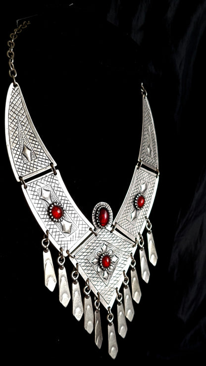 side view of A silver necklace with red stones on a black background. The necklace is made of silver and has a delicate design. The red stones are round and have a deep red color. The necklace is long and hangs down to the chest.