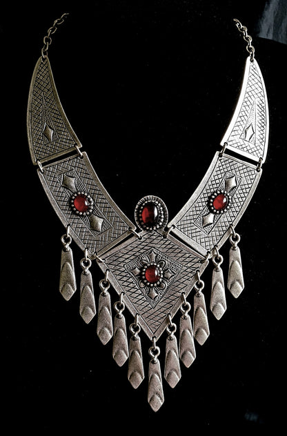 A silver necklace with red stones on a black background. The necklace is made of silver and has a delicate design. The red stones are round and have a deep red color. The necklace is long and hangs down to the chest.