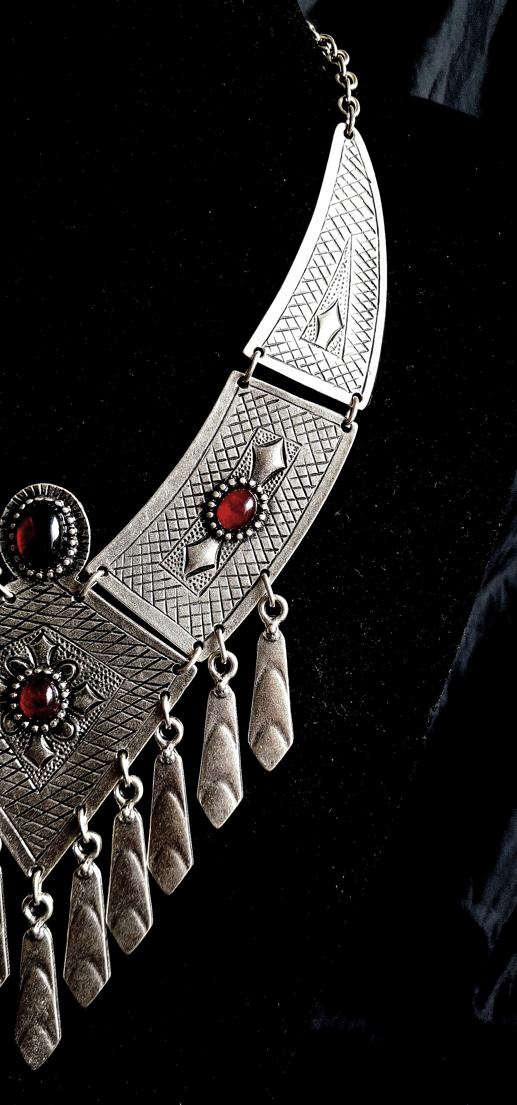 close up view of A silver necklace with red stones on a black background. The necklace is made of silver and has a delicate design. The red stones are round and have a deep red color. The necklace is long and hangs down to the chest.