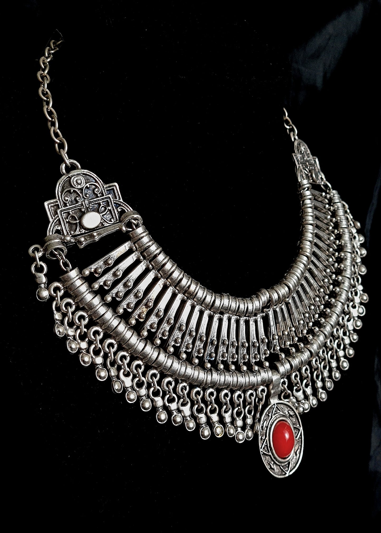 side view of A silver and red necklace on a black background. The necklace is a long, filigree chain with a drop-shaped pendant. The pendant is made of silver and has a red stone in the center. The stone is a deep red color and has a polished finish. The background is a black velvet cloth.