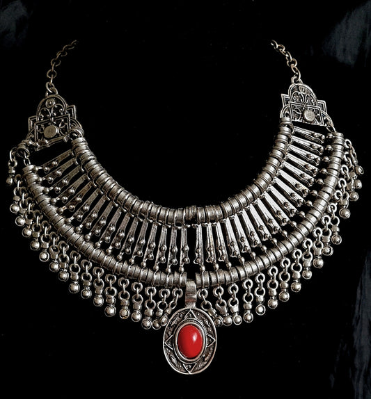 A silver and red necklace on a black background. The necklace is a long, filigree chain with a drop-shaped pendant. The pendant is made of silver and has a red stone in the center. The stone is a deep red color and has a polished finish. The background is a black velvet cloth.