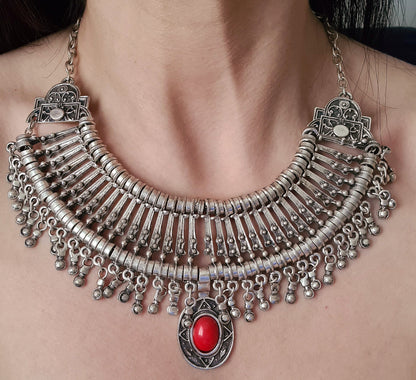 A woman wearing a silver necklace with a red stone in the center. The necklace is a long, filigree chain with a drop-shaped pendant. The stone is a deep red color and has a polished finish.