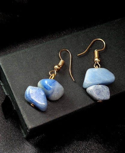 Pair of blue earrings with gold hooks. The earrings are sitting on a black box. The earrings are made of blue rocks and gold hooks. The rocks are oval-shaped and have a smooth surface. The earrings are a beautiful and stylish accessory that would be perfect for any occasion.