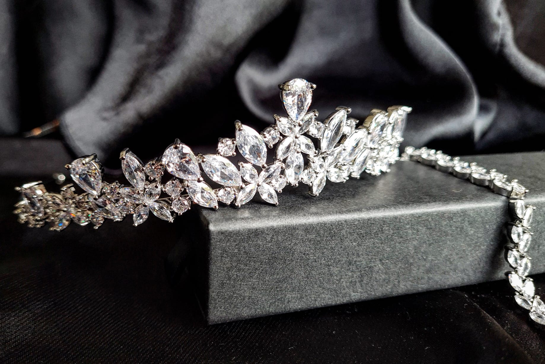 A tiara with diamonds on a black background. The tiara is made of silver and features a delicate design with cascading diamonds. The diamonds are clear and sparkling. The tiara is sitting on a table.