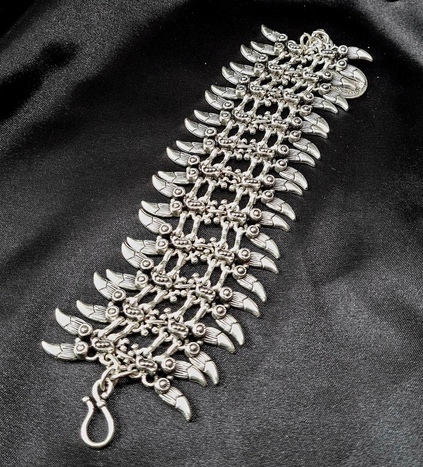 A silver bracelet with a claw design on it. The bracelet is made of silver and has a delicate design. The claw design is in the center of the bracelet and is made up of small, interlocking pieces. The bracelet is sitting on a black cloth