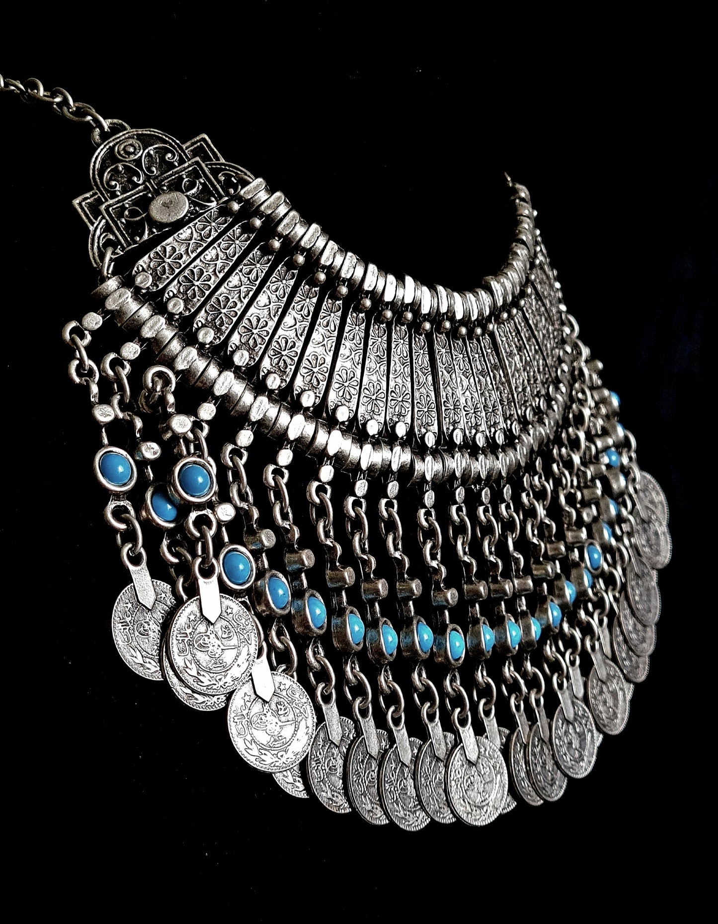 side view of A silver necklace with turquoise beads and coins on a black background. The necklace is made of silver and has a delicate design. The turquoise beads are small and round, and they have a bright blue color. The coins are also small and round, and they have a bronze color. The necklace is long and hangs down to the chest.