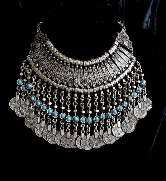 A silver necklace with turquoise beads and coins on a black background. The necklace is made of silver and has a delicate design. The turquoise beads are small and round, and they have a bright blue color. The coins are also small and round, and they have a bronze color. The necklace is long and hangs down to the chest.
