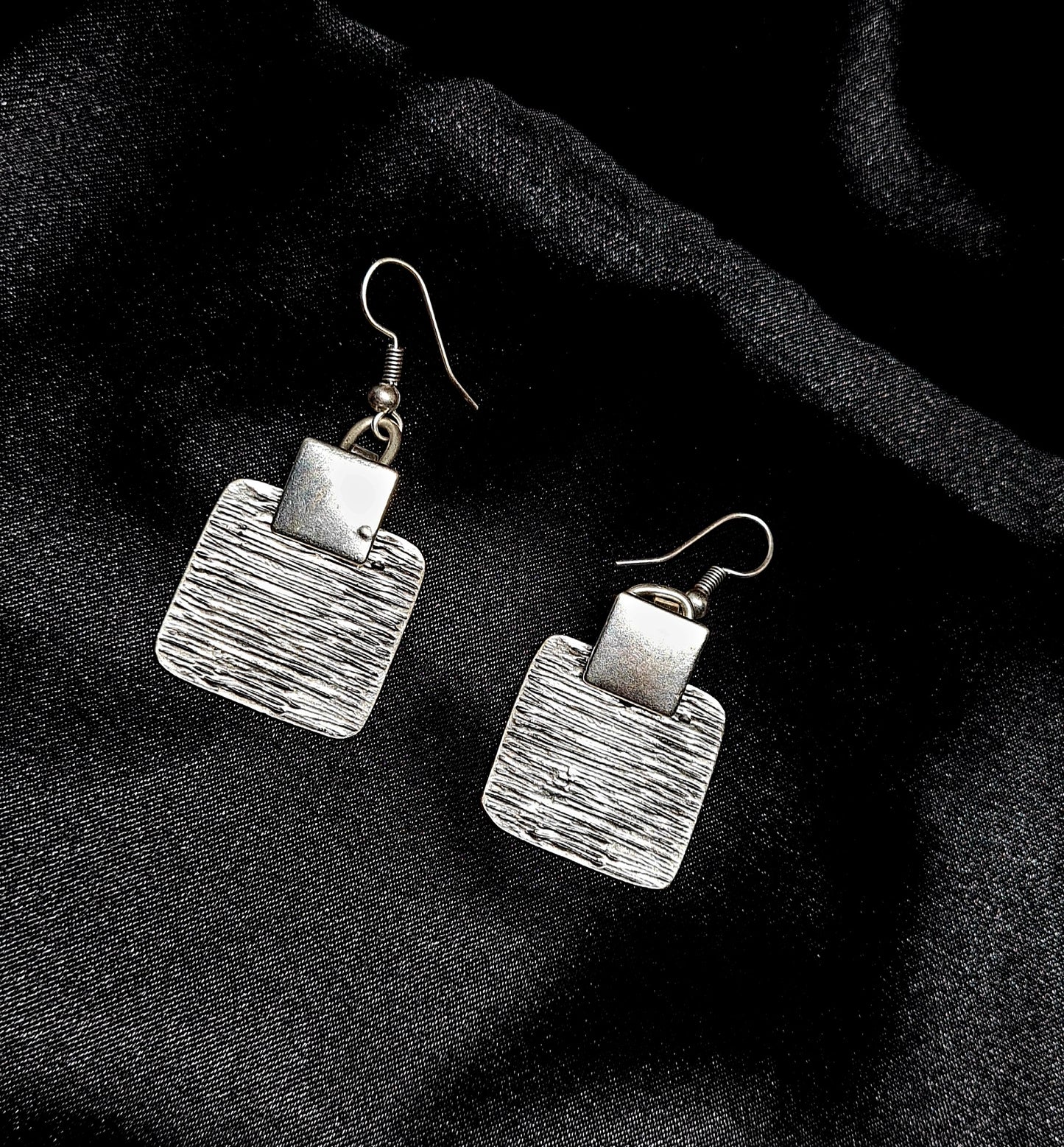 Pair of square sterling silver earrings with a fine linear texture, presented on a minimalist deep black background with soft rim lighting.