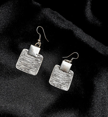 Pair of square sterling silver earrings with a fine linear texture, presented on a minimalist deep black background with soft rim lighting.